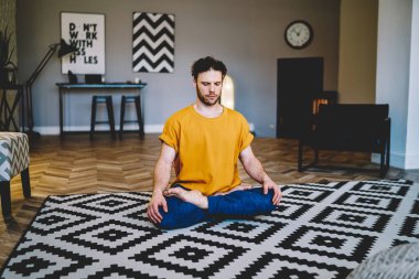 Tüylü genç adam yoga minderinde oturuyor, gözleri modern oturma odasının ortasında kapalı ve rahat koltukta dinleniyor.