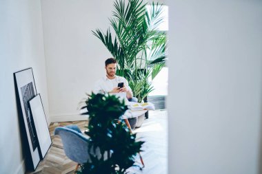 Neşeli, sakallı, gözlüklü bir erkeğin cep telefonuyla masaya oturmuş gündüz vakti büyük bitkiler arasında yaratıcı bir alanda kitap okuması.