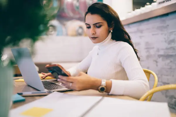 stock image Concentrated serious brunette female in casual white clothes checking information with mobile phone and laptop while doing homework in cafeteria at daytime