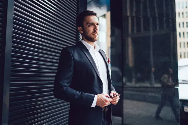 Serious smart businessman in dark suit with mobile phone  standing nearby metal wall and waiting looking away in downtown