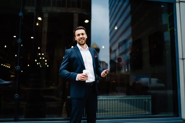 Cheerful relaxed businessman in dark suit with mobile phone  and coffee to go standing on background of window reflecting city lights and looking at camera in downtown