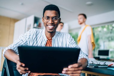 Sıradan giyinmiş, gözlüklü, tablet sörf yapan sosyal medyada kameraya bakan neşeli Afro-Amerikan adamdan.