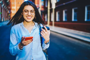 Güneşli bir günde sokakta yürürken, elinde akıllı telefon, elinde mutlu genç hippi kız şehirdeki 4G 'ye bağlı cep telefonundan mesajları kontrol ediyor.