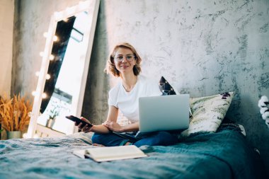 Optik gözlüklü güzel bir kadının portresi kameraya bakıyor ve akıllı telefon ve dizüstü bilgisayarla çevrimiçi mesajlaşmak için boş günlerinde gülümsüyor.