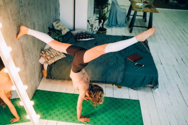 From above athletic adult female in sportswear exercising yoga asanas on green rug in handstand with arm balance and in room with modern bed at home  clipart