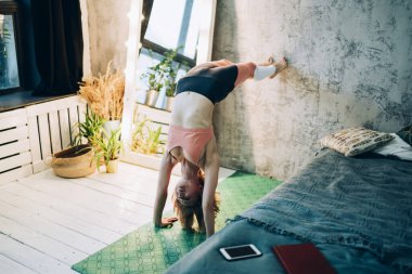 From above athletic adult female in sportswear exercising yoga asanas on green rug in scorpion handstand vrschikasana ii pose in room with modern bed at home  clipart