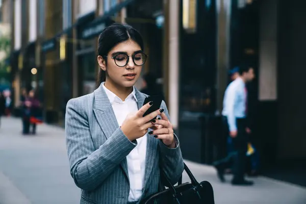 Serious professional expert of exchange standing at urban setting with mobile phone and typing text strategy,Hispanic woman with cellphone gadget waiting for entrepreneur partner outdoors checking sms