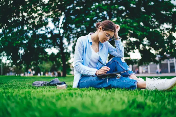 Young creative designer enjoying work outdoors on green grass making sketch in notepad, serious woman student preparing for homework in college campus writing articles concentrated on project