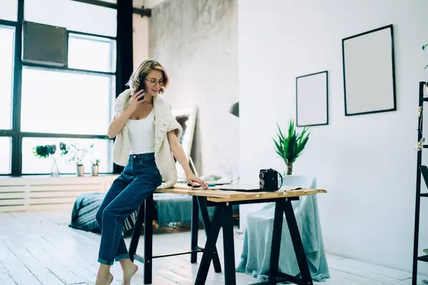 Pretty joyful woman sitting on edge of wooden table barefoot in middle of large bright room and speaking on phone