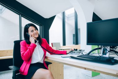 Resmi destek hizmetlerinde çalışan gülümseyen kadın operatörün portresi telefonla olumlu bir konuşma yapıyor, Kafkasyalı iş kadını telefonu ahizesini bilgisayarla görüşmek için kullanıyor.