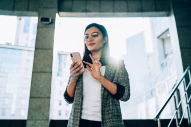 Beyaz kadın milenyum yazı yazma pozitifliği, akıllı telefonlarda şehir planında duruyor, göz giyimli çekici modacı kız sokakta sosyal ağlarda sosyal ağlarda blog yazıyor.