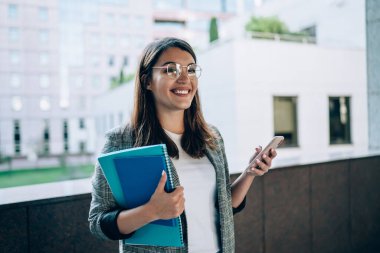 Gözlüklü, resmi kıyafetleri olan neşeli koyu renk saçlı öğrenci modern binanın yanında duruyor ve şehirdeki akıllı telefonla iletişim kuruyor.