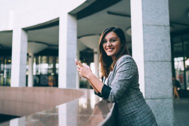 Gözlüklü, modern akıllı telefon ve uygulamalar kullanarak kameraya bakan neşeli, beyaz tenli hippi kız. Cep telefonuyla alışveriş yapmaktan memnun olan mutlu genç kadın. 