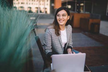 Gözlüklü neşeli, zeki bir kadın dizüstü bilgisayarını karıştırırken ve şehir sokaklarının bulanık arka planında uzaktan iş yaparken başka tarafa bakıyor ve düşünüyor.