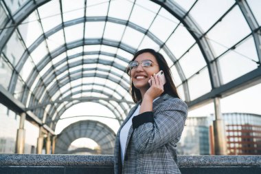 Modern şehrin kemerli geçidinde dururken telefona cevap verirken gülümseyen ve başka yöne bakan gözlüklü neşeli genç bayanın alçak açısı.