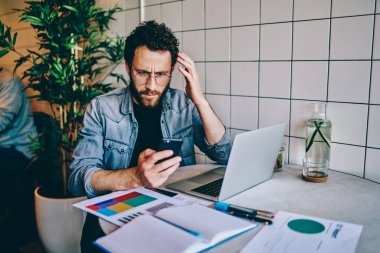 Kafası karışmış, gözlüklü, kafede dizüstü bilgisayarla otururken cep telefonundan beklenmedik iş haberleri okuyan yetişkin bir adam.