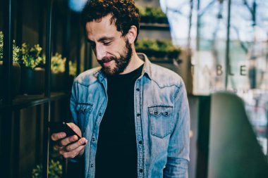 Modern bir kafede dururken, mutlu siyah sakallı şık kıyafetli, gülümseyen ve mesajlaşan bir adam. 