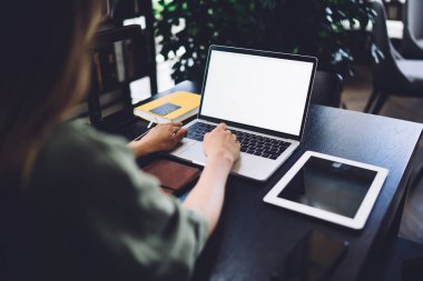 Boş beyaz ekranlı dizüstü bilgisayarda çalışan ve iş yerinde tablet ve defterle masada oturan yüzü olmayan kadının arka planı.