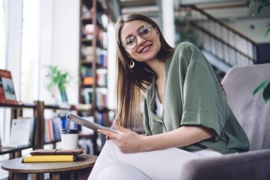 Modern kütüphanede oturup kameraya bakarken, serbest çalışmak için tablet kullanan uzaktan kumandalı genç bayanın yan görüntüsü