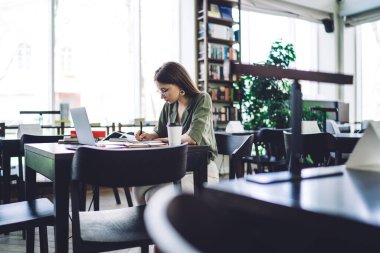 Kitap ve dizüstü bilgisayarla kütüphanede otururken, uzun saçlı, zeki bir kadının not defterine not almasının yan görüntüsü