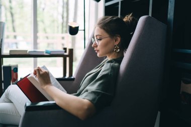 Huzurlu bir kütüphanede rahatlarken rahat koltukta oturup kitap okuyan güzel bir kadın.