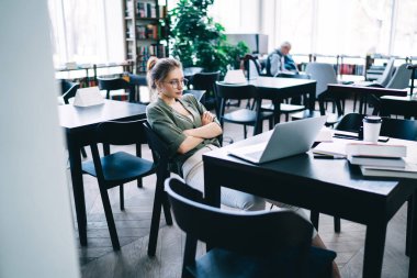 Kitaplarla çevrili bir masada dizüstü bilgisayara bakan ve ışık kütüphanesinde çapraz kollarla düşünen ciddi yorgun bir öğrenci kadının yan görüntüsü.