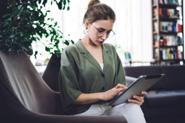 Gözlüklü zeki bir kız büyük boş bir kütüphanede düşünceli yüzüyle ekranda dokunurken tablet üzerinde çalışıyor.