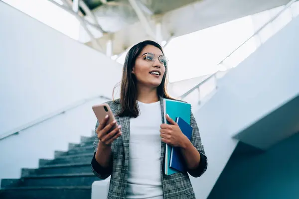Low angle of modern young woman in glasses and formal clothes with notepads looking away while standing on stairs and using smartphone in modern building