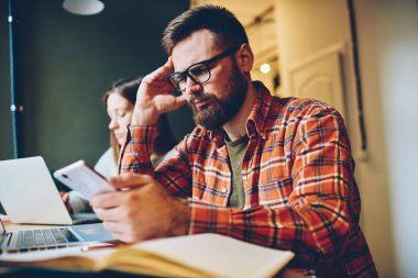 Gözlüklü ve kareli gömlekli stresli adam akıllı telefon kullanıyor ve kafeteryada kadın iş arkadaşının yanında oturup uzaktan kumandayla iş yaparken düşünüyor.