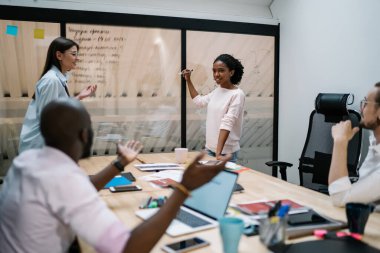 Afro-Amerikalı, günlük kıyafetlerle meşgul bir kadın. Cam tahtaya resim çizen meslektaşlarıyla birlikte ofis alanındaki çalışma planını açıklayan bir proje yaratıyor.