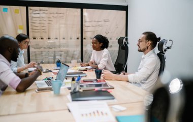 Gündelik giysiler içinde kendinden emin Afrikalı Amerikalı bir kadın modern ofiste masa başında oturan iş arkadaşlarıyla kooperatif bir başlangıç yaratıyor.