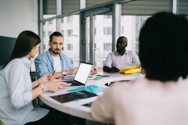 Çok ırklı iş adamları resmi giysiler içinde dairesel masada oturmuş iş stratejisi ve ofis fikirleri hakkında konuşuyorlar.