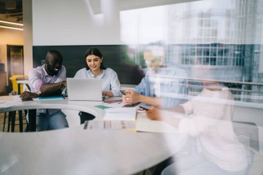 Ofiste birlikte çalışmak için cep telefonu ve dizüstü bilgisayar kullanan genç çalışanların çok ırklı çalışma grubu cam duvarlara yansıyan en yakın binalar