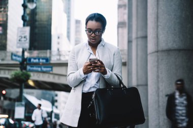 Modern şehir sokaklarının bulanık arka planında dururken sosyal medyada mesaj atan şık çantalı odaklanmış Afrikalı Amerikalı kadın.