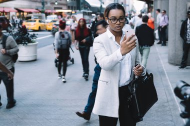 Şık giyinmiş, gözlüklü ciddi bir siyahi kadın. Kalabalık bir caddede dikilirken akıllı telefondan görüntülü sohbet uygulaması kullanıyor.