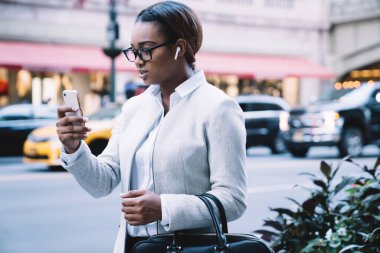 Beyaz takım elbiseli, kablosuz kulaklıklı düşünceli Afro-Amerikan kadın. Sokakta yürürken cep telefonuna dikkatle bakıyor. 