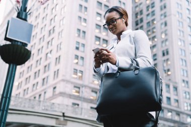 Başarılı Afrikalı Amerikalı iş kadını şık resmi takım elbiseli, arka planda gökdelenlerle şehir caddesinde dikilirken akıllı telefondan mesajlaşıyor.