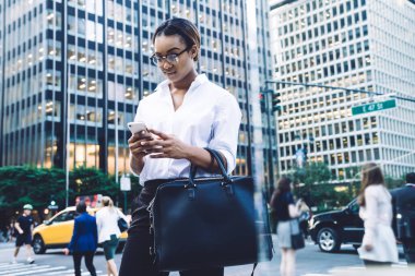 Gözlüklü, beyaz bluzlu, cep telefonuyla mesajlaşan ve kalabalık şehir sokaklarında duran ciddi Afro-Amerikan kadın çalışanı.