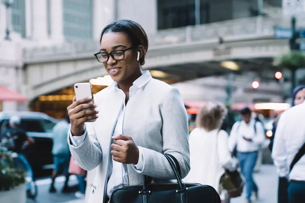 Pleasant African American woman in white classic suit and glasses with wireless earphone in ear talking on mobile phone in street full of people