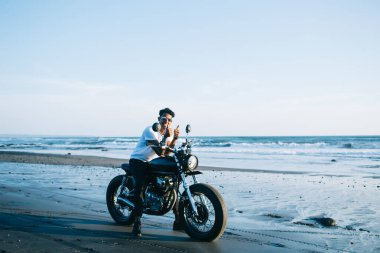 Güneş gözlüklü, pozitif stil sahibi bir adam. Mavi gökyüzü üzerinde dalgalı bir sahil kenarında boş kumlu sahilde park etmiş motosiklette otururken baş parmağını kaldırıp gülümsüyor.