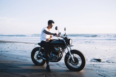 Günbatımında temiz hava ve esintinin tadını çıkarırken motosikletle boş kumsalda tek başına oturan yakışıklı etnik adam.