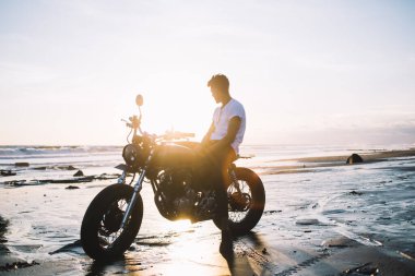 Sıradan giysiler içinde tanınmayan bir adamın motosiklette otururken ve açık havada güneş doğarken ıslak okyanus kumsalına bakarken manzaranın keyfini çıkarması.