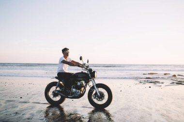 Motosikletli, günlük kıyafetler ve şık güneş gözlüklü, boş deniz kıyısında gezerken eğlenen etnik sürücünün yan görüntüsü.