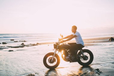 Günbatımında deniz kenarında motosiklet sürerken günlük kıyafetlerle özgürlüğün ve aktif yaşam tarzının tadını çıkaran genç erkek motosikletçinin yan görüntüsü.