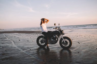 Güneş gözlüklü genç motosikletlinin yandan görünüşü ellerini başının arkasına koyması ve yazın deniz kenarında motosiklette otururken günbatımının keyfini çıkarması.