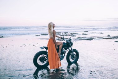 Boş okyanus kıyısında motosiklet üzerinde otururken deniz meltemiyle keyif alan huzurlu sarışın kadın tatilcinin yan görüntüsü. 