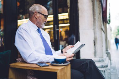 Beyaz gömlekli, kahve fincanı olan yaşlı, kısa saçlı bir adamın şehrin caddesinde pencere ve yol kenarında oturup gazete okumasının yan görüntüsü.