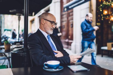 Takım elbiseli ve gözlüklü yaşlı kel bir adamın sokak kafesinde oturup bulanık şehir geçmişi hakkında gazete okumasının yan görüntüsü. 