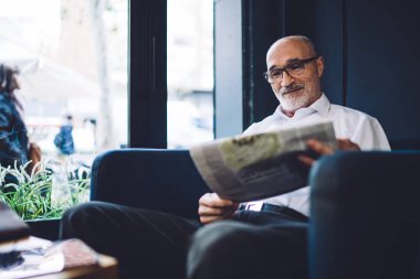 Mutlu kel, gri sakallı, gözlüklü, beyaz gömlekli, pencerenin yanındaki mavi sandalyede oturan ve gazete okuyan yaşlı bir erkek. 