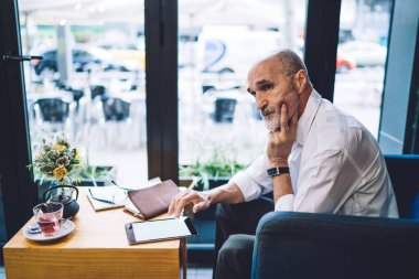 Beyaz gömlekli ve beyaz sakallı sörf tabletli yaşlı işadamının yan görünüşü. Not defteri ve bir fincan çayla masaya oturmuş pencereden dışarı bakan güneş ışığının arka planında el ele yanağa otururken. 